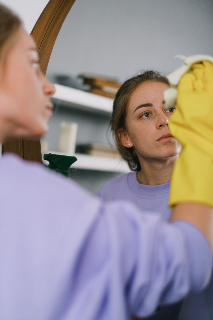 services-02 Female crop in a purple sweatshirt and latex gloves cleaning a mirror in a bright apartment during the day